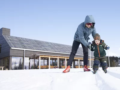 Et bilde av en mamma som hjelper barnet sitt med å gå langrenn, med gravsetstugu i bakgrunnen.