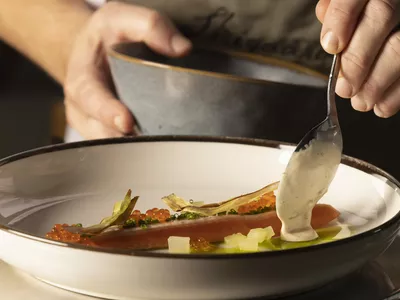 Fiskerett med delikat topping, og en hånd som bruker en skje til å helle saus.