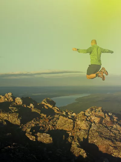 En mann som hopper på toppen av et fjell, han er ikledd grønnjakke.