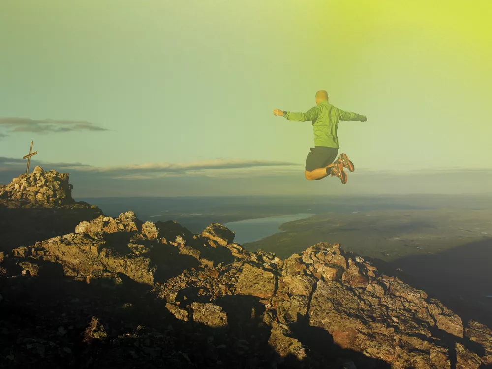 En mann som hopper på toppen av et fjell, han er ikledd grønnjakke.